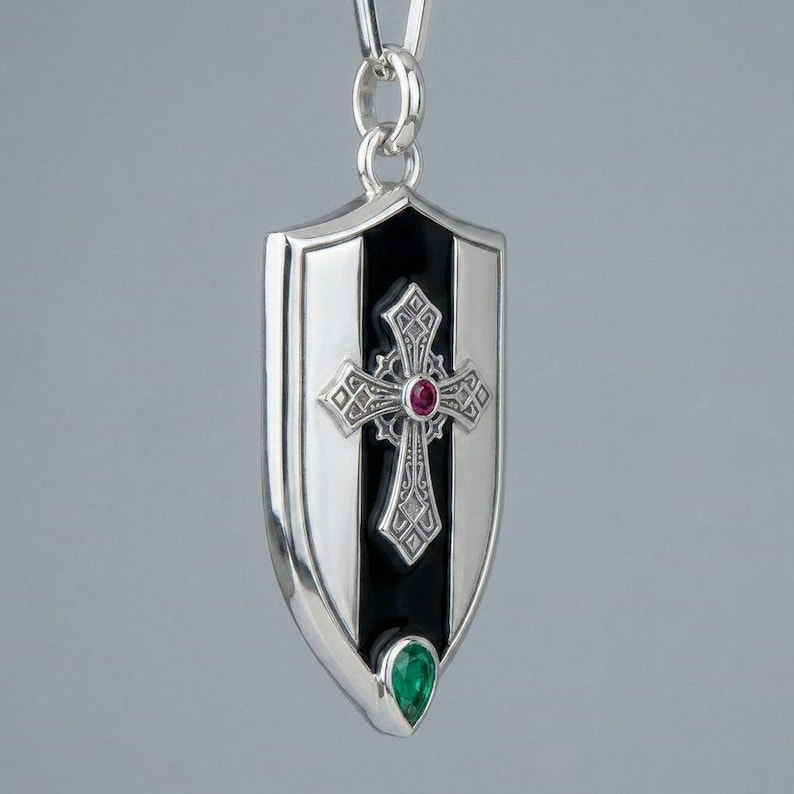 May include: A silver pendant shaped like a shield, featuring a central black vertical stripe with an ornate silver cross overlay. The cross is accented with a small round red gemstone at its centre, and a teardrop-shaped green gemstone sits below.
