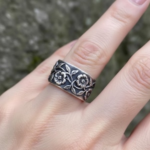Pu&ograve; includere: Un anello d'argento con un motivo floreale. L'anello presenta intagli dettagliati di fiori e foglie attorno alla fascia. L'anello &egrave; indossato su un dito, mettendo in risalto l'intricata lavorazione artigianale e l'estetica generale dell'anello.