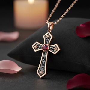May include: A rose gold cross pendant on a matching chain, featuring a dark oxidized silver inlay with intricate vine and rose motifs. A single round, faceted red gemstone is set at the center of the cross, which has polished rose gold edges.