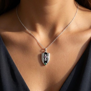 May include: A silver necklace featuring a shield-shaped pendant with a black enamel centre. The pendant displays an ornate silver cross with a small red gemstone at its centre and a single green gemstone positioned at the base of the shield.