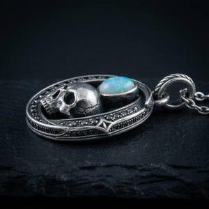 Peut inclure: Un pendentif en argent compos&eacute; d'un cadre ovale incrust&eacute; de petites pierres noires. &Agrave; l'int&eacute;rieur du cadre se trouve un cr&acirc;ne en argent d&eacute;taill&eacute; &agrave; c&ocirc;t&eacute; d'un cabochon d'opale ovale, lisse et iris&eacute;. Le pendentif est suspendu &agrave; une cha&icirc;ne en argent.