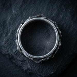 May include: A silver-toned ring featuring a textured, uneven outer band with a dark, oxidized finish. The interior of the band is smooth and polished, contrasting with the rugged, branch-like appearance of the exterior surface of the ring.