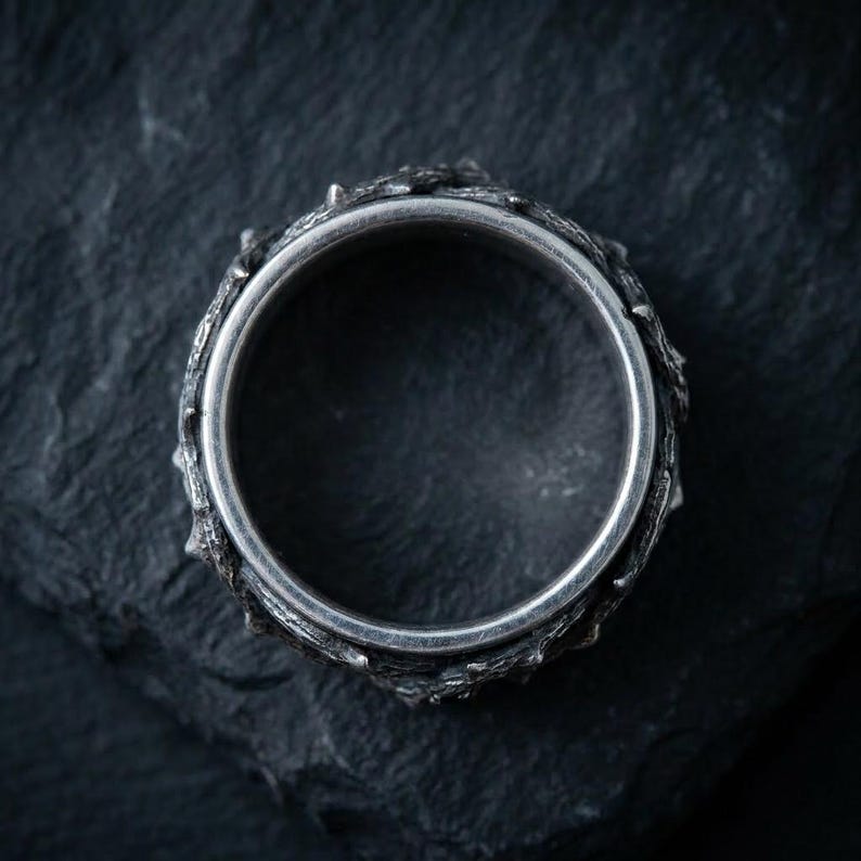 May include: A silver-toned ring featuring a textured, uneven outer band with a dark, oxidized finish. The interior of the band is smooth and polished, contrasting with the rugged, branch-like appearance of the exterior surface of the ring.