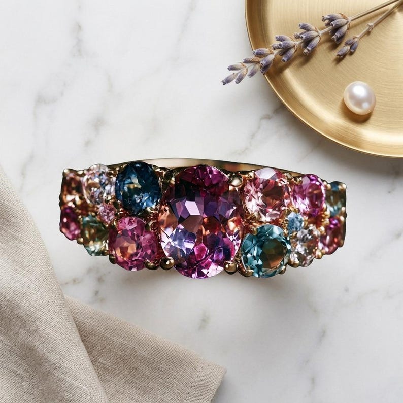 Puede incluir: Un anillo de oro con una gran piedra preciosa central de corte ovalado en color p&uacute;rpura, rodeada por un grupo de piedras m&aacute;s peque&ntilde;as de corte redondo en tonos rosa, azul y verde. El aro est&aacute; hecho de oro pulido con gemas facetadas.