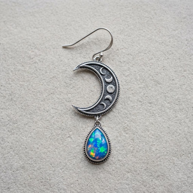 May include: A silver-toned earring featuring a crescent moon charm with engraved lunar phases and a dangling teardrop-shaped stone with iridescent blue and green flashes. The piece includes a curved ear wire for attachment to the earlobe.