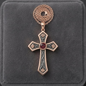 May include: A rose gold cross pendant necklace featuring a central round red gemstone. The cross has a dark grey textured background with rose gold rose motifs at the ends and a rose gold chain coiled neatly above the pendant.
