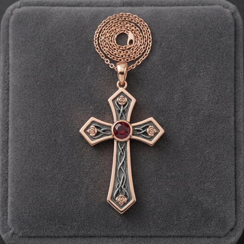 May include: A rose gold cross pendant necklace featuring a central round red gemstone. The cross has a dark grey textured background with rose gold rose motifs at the ends and a rose gold chain coiled neatly above the pendant.