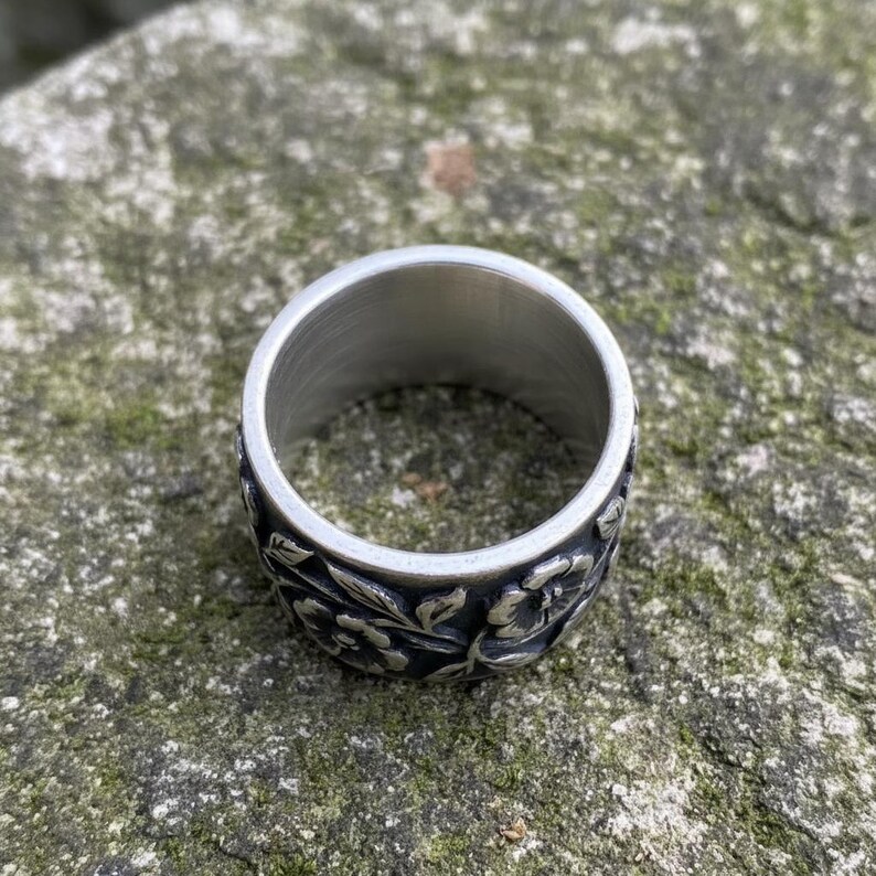 Pu&ograve; includere: Un anello color argento con un motivo floreale. La fascia larga dell'anello presenta un motivo dettagliato di fiori e foglie. L'anello &egrave; realizzato in un materiale metallico lucido.