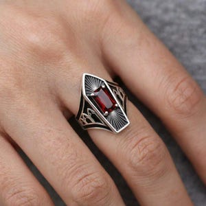 May include: A silver ring featuring a rectangular, deep red gemstone set in a coffin-shaped frame. The metal band displays intricate, dark-oxidized engraved details, including sunburst patterns above and below the stone and arched motifs on the sides.