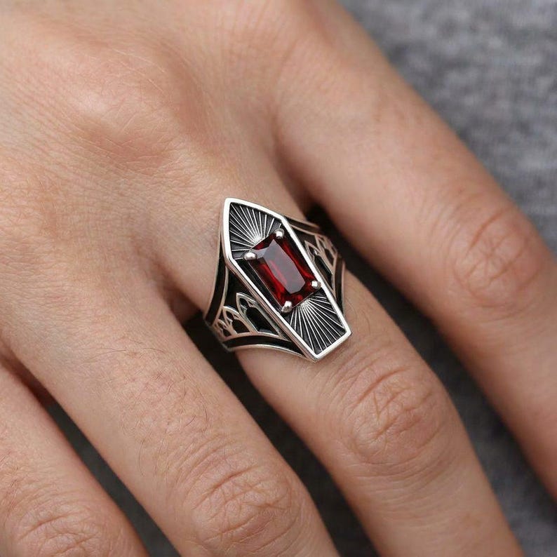 May include: A silver ring featuring a rectangular, deep red gemstone set in a coffin-shaped frame. The metal band displays intricate, dark-oxidized engraved details, including sunburst patterns above and below the stone and arched motifs on the sides.