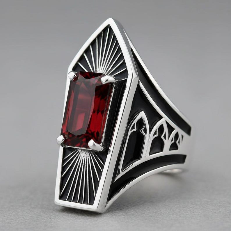May include: A silver ring featuring a rectangular, deep red gemstone set in a coffin-shaped frame. The metalwork includes black enamel accents, sunburst engravings above and below the stone, and arched window cutouts along the side of the band.