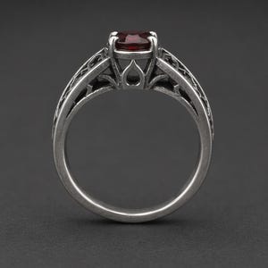 May include: A silver-toned ring featuring a round, deep red gemstone set in a prong setting. The band displays an intricate, openwork filigree pattern with a darkened, oxidised finish that highlights the detailed metalwork throughout the design.