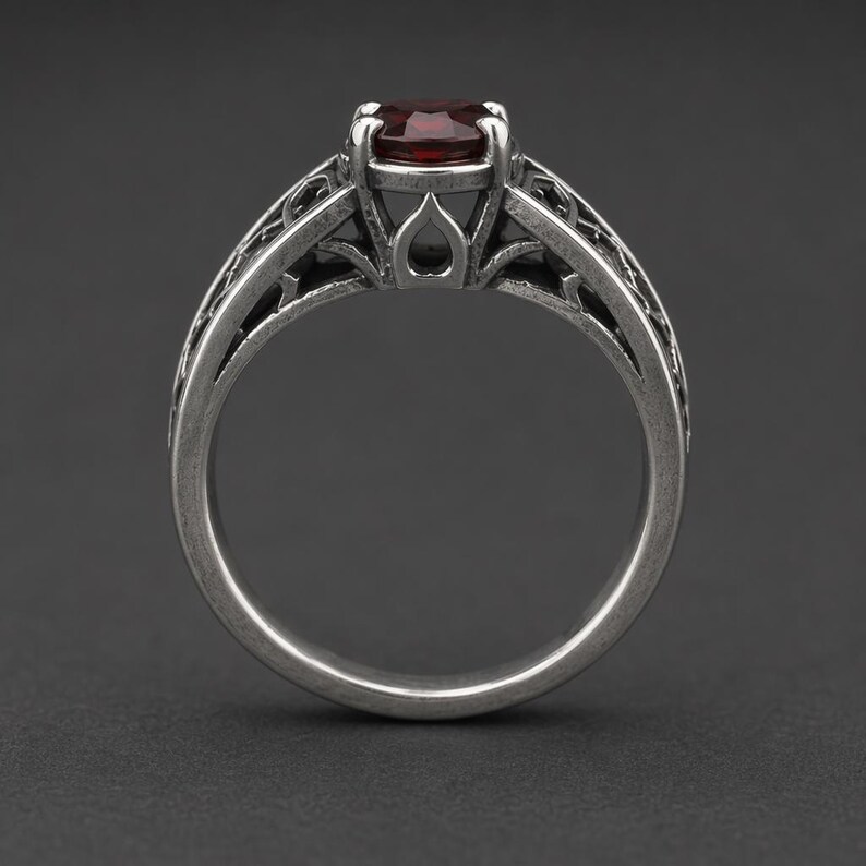 May include: A silver-toned ring featuring a round, deep red gemstone set in a prong setting. The band displays an intricate, openwork filigree pattern with a darkened, oxidised finish that highlights the detailed metalwork throughout the design.