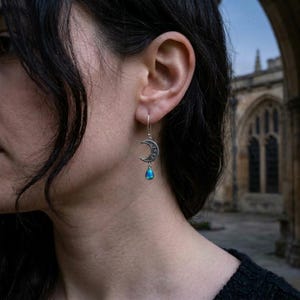 May include: A silver-toned earring featuring a crescent moon charm with engraved lunar phases and a small, iridescent blue teardrop-shaped stone dangling from the bottom, suspended from a simple silver hook wire.
