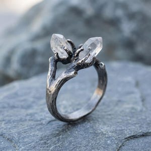 Peut inclure: Bague en argent avec un motif de branche tenant deux pointes de cristal de quartz transparent. La bague a un aspect textur&eacute; et rustique, les cristaux &eacute;tant maintenus en place par les branches. Fond gris.
