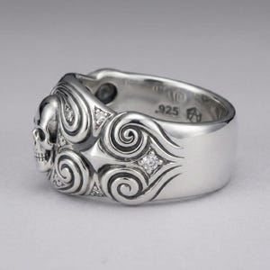 K&ouml;nnte beinhalten: Ein polierter Silberring mit einem skulptierten Totenkopf auf einer Seite, umgeben von komplizierten Wirbelmustern und kleinen, eingelassenen klaren Steinen. Die Innenseite des Rings ist deutlich mit dem Stempel .925 f&uuml;r Sterlingsilber versehen.