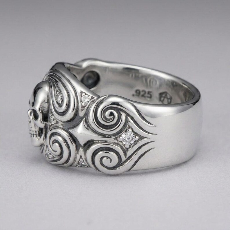 K&ouml;nnte beinhalten: Ein polierter Silberring mit einem skulptierten Totenkopf auf einer Seite, umgeben von komplizierten Wirbelmustern und kleinen, eingelassenen klaren Steinen. Die Innenseite des Rings ist deutlich mit dem Stempel .925 f&uuml;r Sterlingsilber versehen.