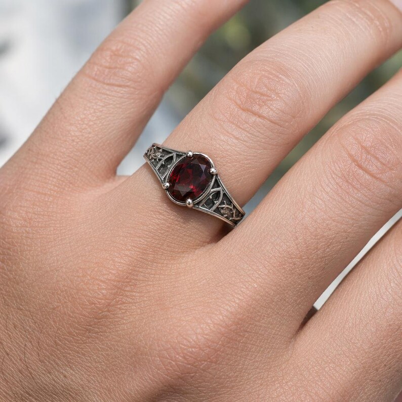 May include: A silver ring featuring an oval-cut deep red garnet gemstone set in a prong setting. The band is crafted with intricate, openwork metal detailing that extends from the central stone along the sides of the ring shank.