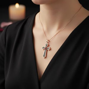 May include: A rose gold chain necklace featuring a cross pendant with a central round red gemstone. The cross has a dark, intricate filigree pattern etched into the metal surface, creating a contrast against the polished rose gold edges.