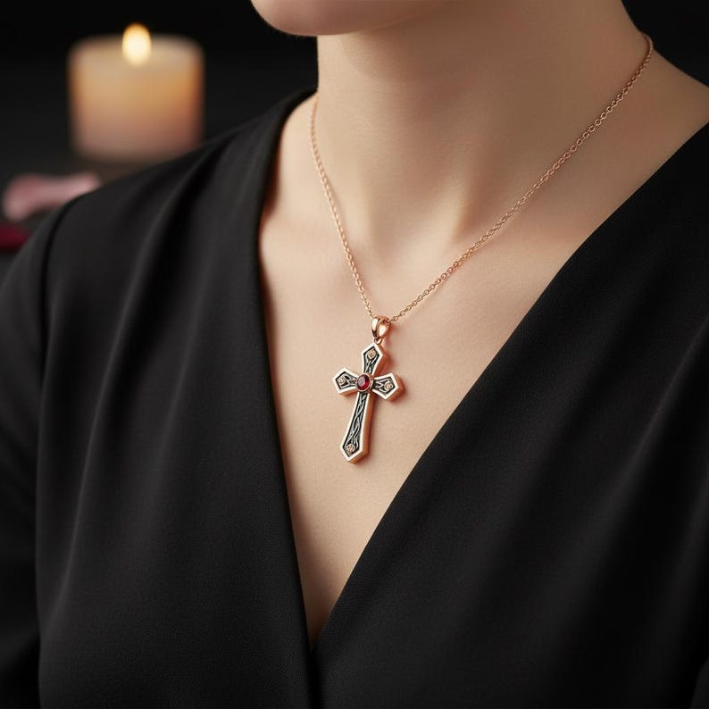 May include: A rose gold chain necklace featuring a cross pendant with a central round red gemstone. The cross has a dark, intricate filigree pattern etched into the metal surface, creating a contrast against the polished rose gold edges.