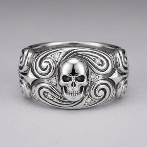K&ouml;nnte beinhalten: Ein silberfarbener Metallring mit einem zentralen Totenkopfmotiv, das von komplizierten Wirbelmustern umgeben ist und mit kleinen, klaren, runden Steinen besetzt ist. Der Ring hat eine polierte Oberfl&auml;che mit symmetrischen sternartigen Formen an den Seiten.