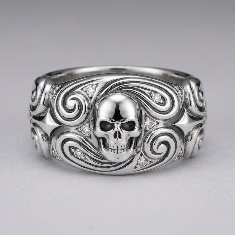 K&ouml;nnte beinhalten: Ein silberfarbener Metallring mit einem zentralen Totenkopfmotiv, das von komplizierten Wirbelmustern umgeben ist und mit kleinen, klaren, runden Steinen besetzt ist. Der Ring hat eine polierte Oberfl&auml;che mit symmetrischen sternartigen Formen an den Seiten.