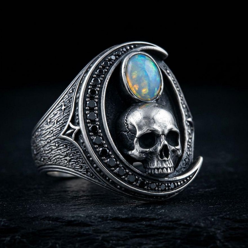 Op de afbeelding: Een zilveren ring in de vorm van een halve maan, versierd met kleine zwarte steentjes. Het midden toont een gedetailleerde menselijke schedel onder een ronde, iriserende opaal cabochon. De ringband heeft een textuur met stervormige gravures.