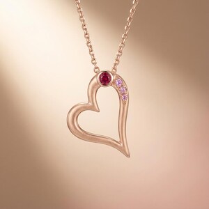 May include: A rose gold heart pendant necklace. The open heart design features a large, round, red gemstone at the top and a cluster of smaller pink gemstones along the curve. The chain is also rose gold.