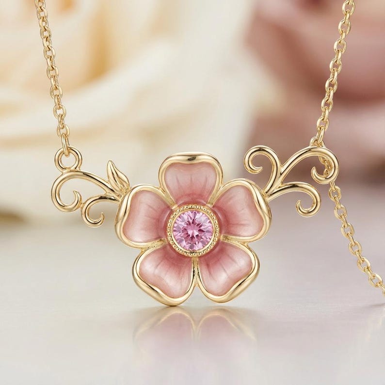 May include: A gold-colored necklace featuring a pink flower pendant with a central pink gemstone. The flower has five petals and is accented with gold-colored swirls and a delicate chain.