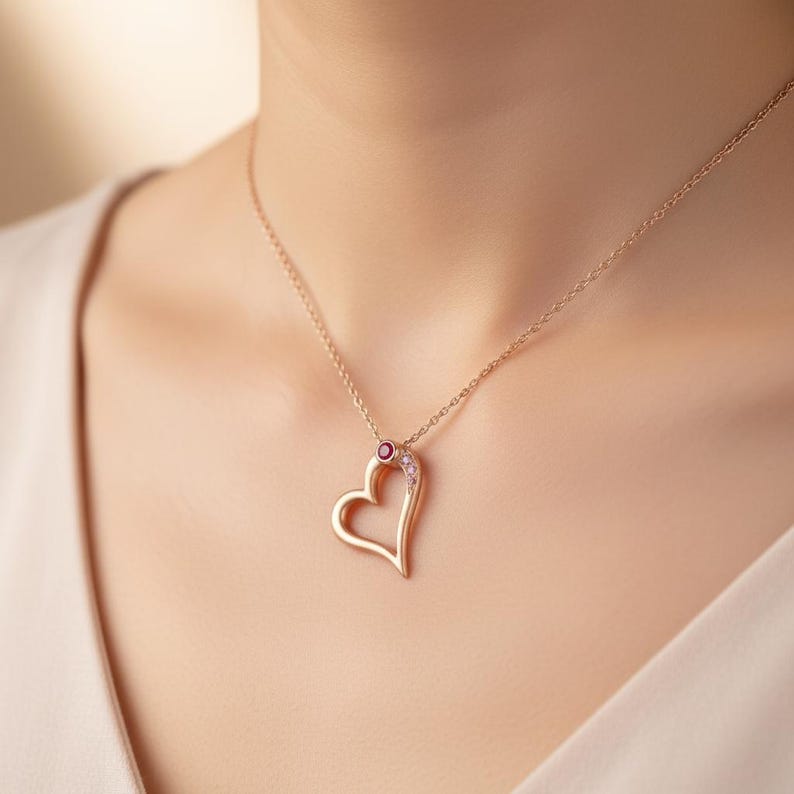 May include: A gold-coloured heart pendant necklace. The open heart design is accented with a small red gemstone and several smaller clear gemstones. The necklace is worn around the neck.