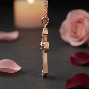 May include: A rose gold cross pendant suspended from a matching delicate chain. The cross features a polished metallic side profile and a small, circular red gemstone set into the center of the cross, visible from this side-angle perspective.