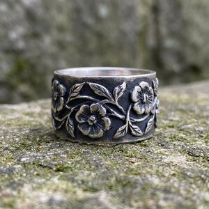 Pu&ograve; includere: Un anello d'argento con un motivo floreale. L'anello presenta incisioni dettagliate di fiori e foglie attorno alla fascia. Ha una finitura scura e ossidata, che gli conferisce un aspetto antico.
