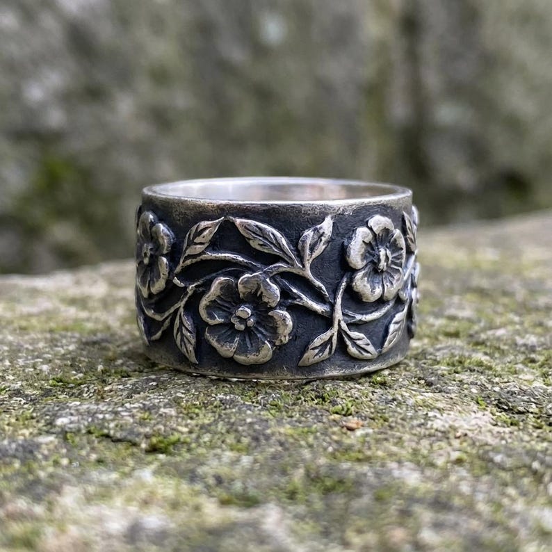 Pu&ograve; includere: Un anello d'argento con un motivo floreale. L'anello presenta incisioni dettagliate di fiori e foglie attorno alla fascia. Ha una finitura scura e ossidata, che gli conferisce un aspetto antico.