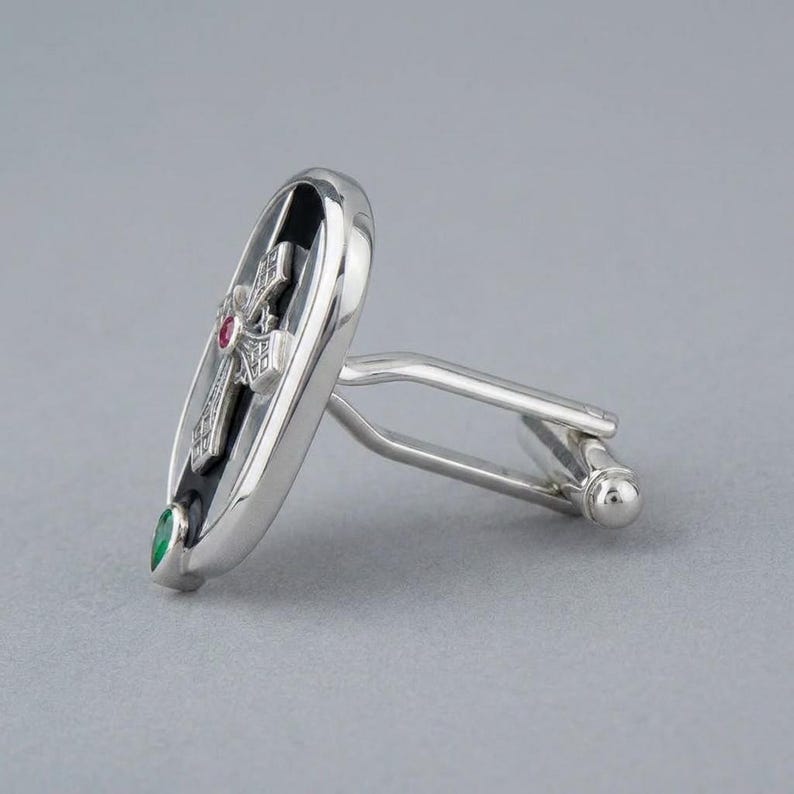 May include: A silver-toned cufflink featuring an oval shape with a black inlay. The centre displays a silver cross motif accented with a small red stone, while a teardrop-shaped green stone is positioned at the bottom of the oval frame.
