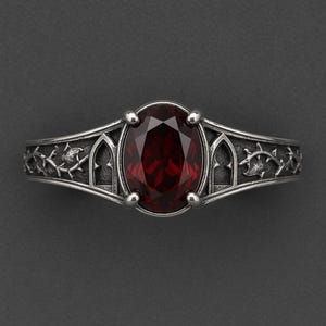 May include: A silver ring featuring a central oval-cut deep red gemstone held by four prongs. The band is decorated with intricate relief carvings of pointed archways and thorny vine patterns, finished with a dark, oxidised metallic texture.