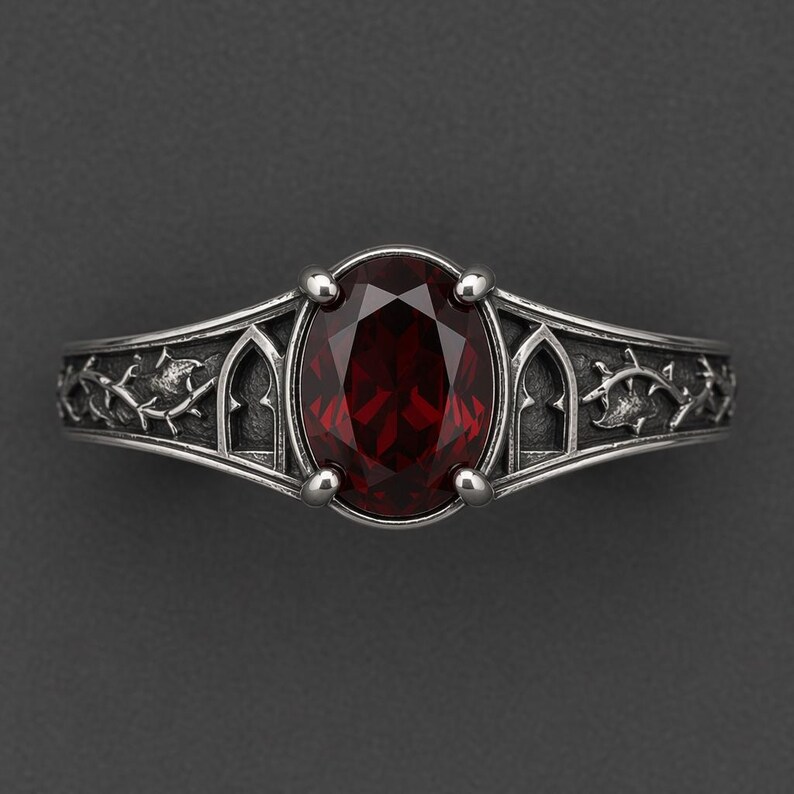 May include: A silver ring featuring a central oval-cut deep red gemstone held by four prongs. The band is decorated with intricate relief carvings of pointed archways and thorny vine patterns, finished with a dark, oxidised metallic texture.