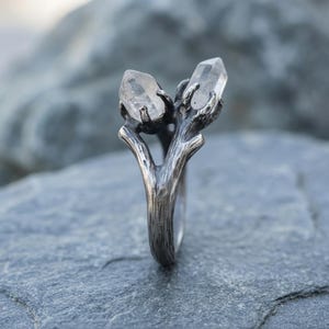 Peut inclure: Bague en argent avec un design en forme de branche tenant deux cristaux de quartz clairs. Les cristaux sont facett&eacute;s et brillent. La bague a une finition textur&eacute;e et antique, lui donnant une esth&eacute;tique rustique et naturelle. Un bijou unique.