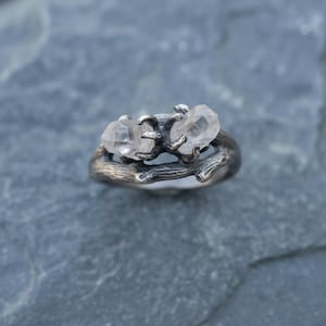 Peut inclure: Une bague en argent orn&eacute;e de deux cristaux de quartz clairs. Le corps de la bague est con&ccedil;u pour ressembler &agrave; une brindille, les cristaux &eacute;tant maintenus par de petites griffes. La bague est pos&eacute;e sur une surface en pierre grise.