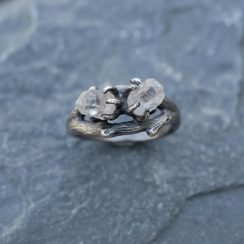 Peut inclure: Une bague en argent orn&eacute;e de deux cristaux de quartz clairs. Le corps de la bague est con&ccedil;u pour ressembler &agrave; une brindille, les cristaux &eacute;tant maintenus par de petites griffes. La bague est pos&eacute;e sur une surface en pierre grise.