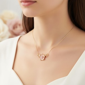 May include: A gold-colored necklace featuring a delicate flower pendant with pink and white petals. The pendant is accented with ornate gold filigree and hangs from a fine chain. The necklace is worn on a person.
