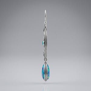 May include: A silver drop earring featuring a circular frame with a beaded edge and a central blue opal inlay. A teardrop-shaped blue opal pendant hangs from the bottom of the circle, suspended by a thin silver hook wire for pierced ears.