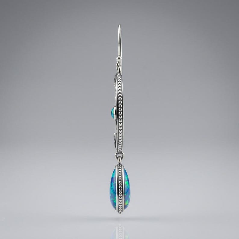 May include: A silver drop earring featuring a circular frame with a beaded edge and a central blue opal inlay. A teardrop-shaped blue opal pendant hangs from the bottom of the circle, suspended by a thin silver hook wire for pierced ears.