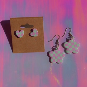 Puede incluir: Un juego de pendientes de cuadros rosa y verde. Un par son tachuelas en forma de corazón y el otro par son pendientes colgantes en forma de flor.