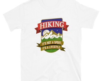 Op de afbeelding: Een wit T-shirt met korte mouwen en een afbeelding van een berglandschap onder een nachtelijke hemel. Een rode banner bovenaan bevat de tekst HIKING, en een groene banner eronder toont IT'S NOT A SPORT, IT'S A LIFESTYLE.