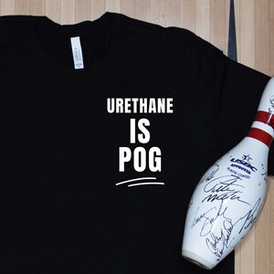 May include: Black t-shirt with the text "URETHANE IS POG" in white. A purple and gold bowling ball and a white bowling pin with signatures are also in the image. The Strike Life logo is at the bottom.
