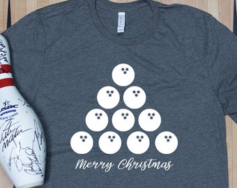May include: A gray t-shirt with a white bowling ball Christmas tree design and the words "Merry Christmas." A bowling pin and a purple bowling ball are also in the image. The shirt is on a wooden surface.