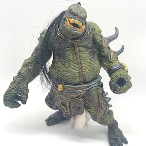 May include: A detailed action figure of a grotesque, green-skinned creature with black hair and yellow teeth. The figure is wearing armor with metallic accents and has a furry tail. The figure is posed in a dynamic stance.