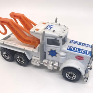 May include: A vintage white and grey toy police tow truck with orange lifting arms. The truck features the word "POLICE" in blue lettering. It has black wheels and a blue star emblem. A classic toy vehicle.
