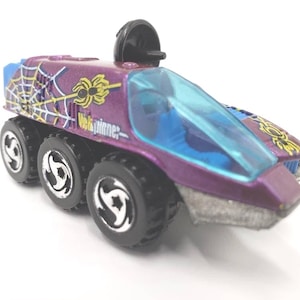 May include: Purple toy car with six black wheels. The car has a blue windshield and a black dome on top. The car is decorated with spider web and spider graphics. The text "Web Spinner" is printed on the side.