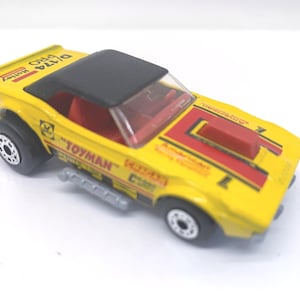 May include: A yellow toy car with a black top and red accents. The car has the word "TOYMAN" printed on the side and "D/174 PRO Holley" on the top. The car has black wheels and a silver exhaust pipe.
